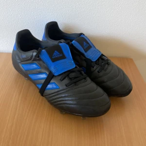 Svarta och blå fotbollsskor från Adidas - Säljer ett par svarta fotbollsskor från Adidas med blå detaljer och klassiska tre ränder på sidan. Skorna har snörning och en bred blå plös med Adidas-logga. Yttermaterialet är syntet och sulan har dobbar för gräsplan. Perfekt för dig som vill ha stil på planen. De är Elite och har används i runt 1 år storlek 40 2/3.      Pris kan diskuteras 