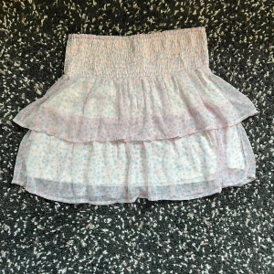 Blommig volangkjol från Vero Moda - Supersöt kortkjol från Vero Moda med blommigt mönster i ljusrosa, blått och lite beige. Kjolen har två lager volanger och en smockad midja som sitter skönt. Små hål men inget som märks(se bild 4) 