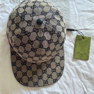Beige keps med GG-mönster från Gucci - Snygg beige keps från Gucci med klassiskt GG-monogram i svart över hela kepsen. Kepsen har en böjd skärm och en röd-blå rand på sidan. Tillverkad i bomullsmix med justerbar rem baktill. Perfekt för dig som vill sticka ut med en lyxig accessoar. 🔥Passa på köpa nu 🔥