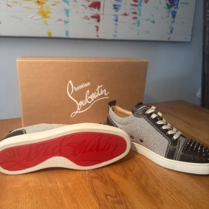 Christian Louboutin sneakers med nitar - Snygga sneakers från Christian Louboutin med signifikant röd sula och coola nitar på tån. Skorna har svart och grå textil med svarta skinnpaneler och vita kontrastsömmar. Klassisk snörning och rund tå. Perfekt för dig som vill sticka ut med exklusiv stil. Nypris ca 10 000, helt nya aldrig använda. 