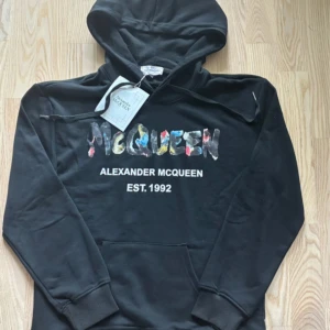 Svart hoodie Alexander McQueen - Svart hoodie från Alexander McQueen med färgglad logotyp framtill och vit text 'EST. 1992'. Tröjan har huva med snörning och en stor magficka. Materialet är mjuk bomull och passformen är normal. Perfekt för dig som gillar streetwear med designer-vibe.