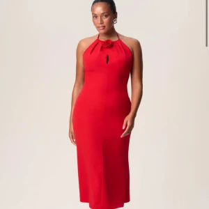 Röd långklänning med rosdetalj - Säljer en elegant röd långklänning med halterneck och en snygg rosdetalj vid halsen. Klänningen har en liten öppning framtill och är figurnära med en stilren siluett. Perfekt för dig som vill sticka ut med en klassisk look och cool detalj vid halsen.