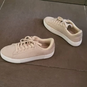 Beige sneakers med vit sula - Snygga beige sneakers med klassisk rund tå och platt vit sula. Skorna har snörning och är tillverkade i mocka-liknande material med mjuk textil på insidan. Perfekt för dig som gillar stilrena och neutrala färger.
