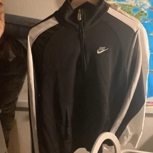 Nike svart och vit vindjacka - Nike vindjacka i svart med vita paneler längs axlar och ärmar. Klassisk sportig look med halv dragkedja och broderad Nike-logga på bröstet. Jackan har hög krage och är tillverkad i ett lätt polyestermaterial, perfekt för en aktiv stil.