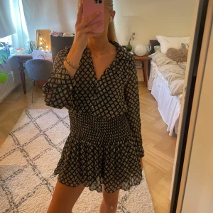 Svartvit kortklänning med mönster - Supersöt kortklänning i svart och vitt med små rutigt mönster. Klänningen har långa ärmar, v-ringning och smockad midja som ger en snygg siluett. Volangig kjol och lätt, luftig känsla. Perfekt för dig som gillar trendiga detaljer! 🩷🩷🩷 Skriv för fler bilder, pris kan diskuteras!!