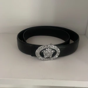 Svart Versace-bälte med silverdetaljer - Snyggt svart bälte från Versace i skinn med en stor, oval silverspänne prydd med Medusa-huvud och glittrande stenar runtom. Bältet har diskret mönsterprägling längs remmen som ger extra lyxig känsla. Perfekt accessoar för att lyfta din outfit.