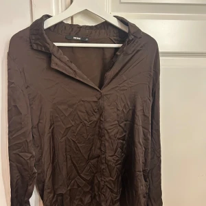 Brun pyjamas skjorta i silkes från Bik Bok  - Snygg brun pyjamas skjorta i silkes från Bik Bok i storlek XS. Skjortan har klassisk krage, knäppning framtill och långa ärmar. Perfekt att styla med jeans eller kjol för en trendig look. Knappt använd