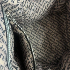 Juicy Couture svart sammetstote - Juicy Couture shoppingväska i svart sammet med breda axelband och innerfoder med logotypmönster. Väskan har en innerficka med dragkedja och medföljande liten silverfärgad plånbok med Juicy Couture-logga i guld. Perfekt för dig som gillar statement-accessoarer.