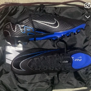 Nike Mercurial svarta/blå fotbollsskor - Snygga Nike Mercurial fotbollsskor i svart med blå detaljer och silverfärgad swoosh. Skorna har snörning och en platt sula med dobbar för bra grepp på planen. Perfekta för dig som vill sticka ut på matchen!