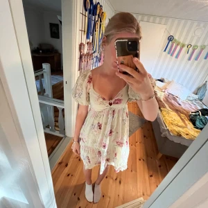 Blommig kortklänning med puffärm - Supersöt kortklänning med ljus botten och rosa blommigt mönster. Klänningen har puffiga kortärmar, v-ringad urringning med liten rosett och volangkant nedtill. Passformen är loose och tyget känns som mjuk bomull, perfekt för sommarens varma dagar.