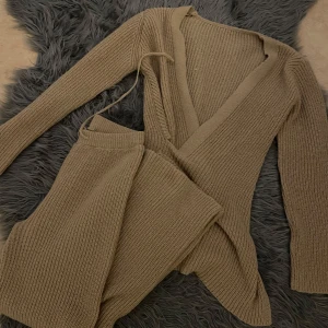 Beige omlott stickad kofta - Säljer en beige stickad kofta med omlott-design och knytband i midjan. Koftan har långärmade och ribbad struktur som ger en mysig och trendig look. Perfekt att slänga på sig när det är lite kyligt ute.kommer med matchande byxor.storlek S på båda