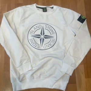Vit Stone Island sweatshirt med logga - Säljer en vit sweatshirt från Stone Island med stor broderad kompasslogga i blått på bröstet och klassisk patch på vänster ärm. Tröjan har rund hals, ribbade muddar och är tillverkad i mjuk bomull. Perfekt för dig som gillar streetwear och snygga detaljer.