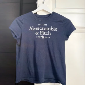 Mörkblå Abercrombie & Fitch t-shirt - Mörkblå t-shirt från Abercrombie & Fitch med vit logotyp och text framtill.