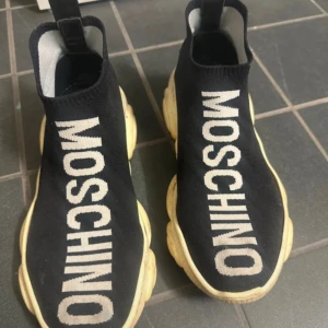 Svarta Moschino sneakers med björnsula - Unika svarta sneakers från Moschino med vit logga längs ovansidan och chunky beige sula formad som en björn. Stickad ovandel i syntetmaterial ger en strump-liknande passform. Perfekta för dig som vill sticka ut med cool design och streetstyle-vibe.