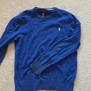 Blå bomullströja från Polo Ralph Lauren - Snygg blå tröja från Polo Ralph Lauren i storlek L. Tröjan är tillverkad i mjuk bomull och har ett klassiskt crewneck-utseende med långa ärmar och ribbade muddar. På bröstet finns den ikoniska vita Polo-loggan broderad. Perfekt för dig som gillar stilren och enkel design.