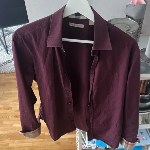 Vinröd Burberry skjorta - Snygg mörklila skjorta från Burberry med klassisk krage och knappar framtill. Ikoniskt rutigt Burberry-tyg på insidan av kragen och broderad logga på bröstet. Tillverkad i mjuk bomull och har långa ärmar med stilrena manschetter.