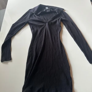 Svart ribbad klänning från H&M Divided - Svart ribbad klänning från H&M Divided i storlek S. Klänningen har lång ärm, v-ringad hals och är figurnära med en kort längd. Materialet är mjukt och stretchigt, perfekt för dig som gillar en slim passform och stilren look.