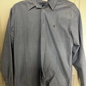 Blå skjorta från Ralph Lauren - Snygg blå skjorta från Ralph Lauren med klassisk button-down krage och broderad logga på bröstet. Skjortan har långa ärmar och knappar framtill. Perfekt för dig som gillar stilrena och tidlösa plagg. Storlek är M/L