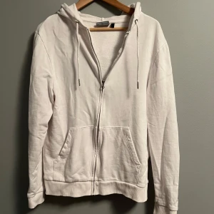 Vit hoodie med dragkedja och huva - Säljer en stilren vit hoodie med dragkedja framtill och två stora fickor. Tröjan har huva med snörning och långa ärmar. Perfekt för chill dagar och enkel att matcha med jeans eller mjukisbyxor. Materialet känns mjukt och bekvämt, troligen bomull eller polyester.