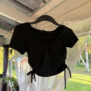 Svart croppad topp med knyt - Svart croppad topp från Shein i storlek XS. T-shirten har korta ärmar, v-ringning och snygga knytband i sidorna som ger en cool detalj. Perfekt till jeans eller kjol för en trendig look.