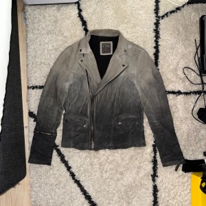 AllSaints Suede Gradient Throttle Jacket - Sällsynt AllSaints mockajacka i unik ljusgrå–svart färggradient. Premium mocka, metall-dragkedja och fina detaljer. Mått: L 60 cm, ärm 64 cm, axel 46 cm, pit to pit 50 cm. Utgången modell som inte säljs längre. I utmärkt skick och byggd för att hålla i många år till.