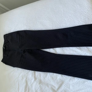 Vilma kostymbyxor  - Snygga svarta kostymbyxor med tunna vita ränder. Byxorna har rak passform och klassisk look, perfekta för en stilren outfit. Materialet känns mjukt och följsamt, och midjan är normal. Passar dig som gillar en tidlös vibe.