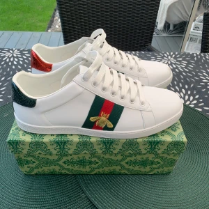 Gucci Ace vita sneakers med bi-detalj - Gucci Ace sneakers i vitt skinn med klassiska gröna och röda ränder på sidorna. Ena sidan har en broderad guldfärgad bi. Jag har använt skorna en gång, skorna är som hell nya, och det ingår allt som fick, skorna är i storlek 43, hoppas dem passar en ny ägare