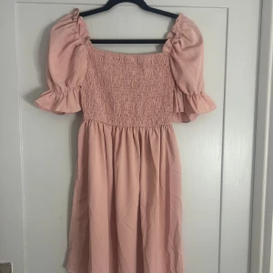 Ljusrosa kortklänning med puffärm - Supersöt ljusrosa klänning med puffiga kortärmar och smockad överdel. Kjolen är lätt utställd och klänningen har en romantisk vibe med volangdetaljer vid ärmslut. Perfekt för dig som gillar en feminin och trendig stil.