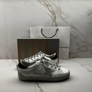 Golden Goose vita sneakers med stjärna - Snygga vita sneakers från Golden Goose med grå stjärndetalj på sidan och svarta detaljer vid hälen. Skorna har klassisk snörning och en något smutsad look som är typisk för märket. Tillverkade i skinn med platt sula och rund tå.