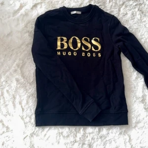 Svart långärmad tröja Hugo Boss - Svart långärmad tröja från Hugo Boss med stor guldfärgad BOSS-logga på bröstet. 
