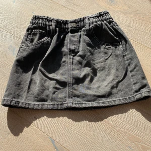 Grå jeanskjol från &Denim, stl 116 - Säljer en grå jeanskjol från &Denim i storlek 116. Kjolen har två bakfickor och en elastisk midja som gör den bekväm och enkel att bära. Klassisk rak modell i mjukt denimtyg, perfekt för dig som gillar enkel och cool stil.