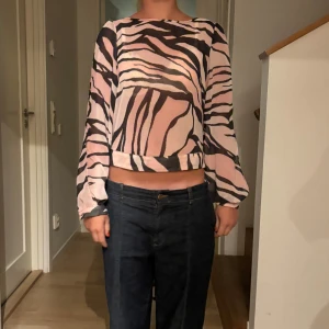 Zebramönstrad croppad blus med öppen rygg - Säljer en luftig och croppad blus med zebramönster i rosa och svart. Toppen har långa ärmar med slits, öppen rygg och öppna armar och knytband bak. Perfekt för dig som vill sticka ut med en cool och trendig look.