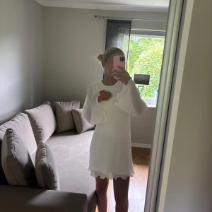 Vit kortklänning med volangärmar - Supersöt vit kortklänning med volangdetaljer och ribbad struktur. Klänningen har långa ärmar med volangkant och rund halsringning. Materialet känns lätt och luftigt, perfekt för ljusa dagar. Stilren och feminin look med extra flow i ärmar och nederkant.