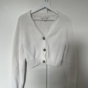 Vit stickad kofta från Monki - Fin vit stickad kofta från Monki i croppad modell. Koftan har v-ringning, långa ärmar och snygga knappar framtill. Perfekt att slänga över en topp för en chill och stilren look.