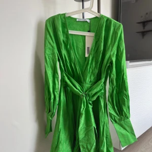 Grön omlottklänning från Adoore - Snygg grön omlottklänning från Adoore i glansigt material. Klänningen har lång ärm med knappar vid manschetten, v-ringad hals och ett brett knytband i midjan med metallring. Perfekt för dig som vill sticka ut med färg och cool siluett.