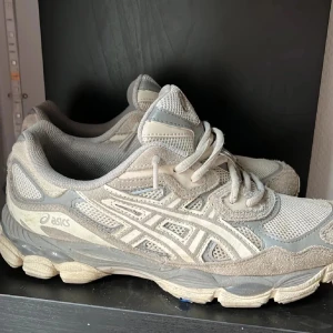 Gråa sneakers från Asics - Säljer ett par gråa Asics sneakers med vit logga och detaljer. Skorna har snörning och en platt sula, tillverkade i en mix av syntet och mocka. Perfekta för dig som gillar sportig stil och vill ha bekväma skor till vardagen.