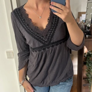 Blus från Odd Molly - Supersnygg grå blus med djup v-ringning och dekorativa spetsdetaljer framtill. Blusen har trekvartsärmar med lätt utsvängd kant och fina spetskanter nertill.