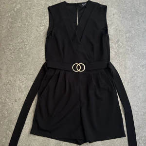 Svart jumpsuit från Sisters Point - Säljer denna snygga svarta jumpsuiten från Sisters Point i kort modell. Jumpsuiten är ärmlös och har ett bälte med guldfärgat spänne i midjan. Tillverkad i mjuk polyester med lite stretch för skön passform. Aldrig använt då jag ej hittat ett bra tillfälle att använda den på.