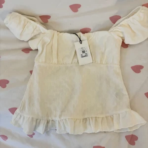 Vit blus med volang och puffärm - Supersöt vit blus med korta puffärmar och volangkant nertill. Toppen har en lätt strukturerad yta och sitter offshoulder för en romantisk vibe. Perfekt till jeans eller kjol för en trendig look.