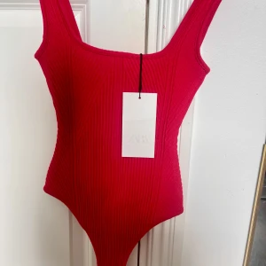 Ribbad röd body från Zara - Ribbad röd body från Zara med breda axelband och fyrkantig ringning. Snygg, tight passform och stretchigt material som sitter figurnära. Säljer då den inte går på mig och jag kan inte lämna tillbaka då jag köpte den i Spanien men lapparna är kvar och den är helt oanvänd. 