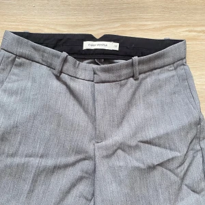 Grå kostymbyxor från Carin Wester - Snygga grå kostymbyxor från Carin Wester i klassisk rak modell. Byxorna har bälteshällor, diskreta fickor och stängs med dragkedja, hake och knapp. Perfekta för en clean och stilren look. Materialet känns mjukt och har en lätt struktur.