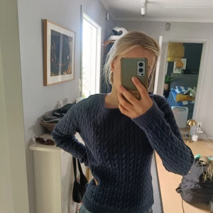 Mörkblå kabelstickad tröja Holly & White - Snygg mörkblå kabelstickad tröja från Holly & White i storlek M. Tröjan har rund hals, ribbade muddar och ett klassiskt flätmönster över hela plagget. Perfekt att slänga på sig när det är lite kyligt ute. Passar dig som gillar enkel och stilren look.