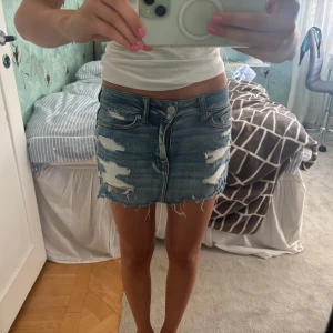 Blå jeanskjol med slitningar - Säljer en kort jeanskjol från Dr. Denim i klassisk blå tvätt med rå kant och slitna detaljer framtill och baktill. Kjolen har fem fickor, knapp och dragkedja samt en cool, fransig finish längst ner. Perfekt för dig som gillar en edgy streetstil. Storlek 32 men passar mig som vanligtvis bär xs/s i underdelar.