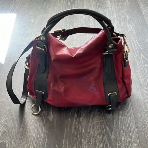 Röd axelväska i skinn med mörk bruna detaljer - Snygg röd axelväska med choklad brun handtag och axelrem. Väskan har guldfärgade metalldetaljer och en rymlig insida med blått foder och flera fack. Perfekt storlek för att få plats med allt du behöver och cool design med kontrastfärger.