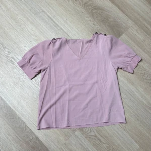 Ljusrosa blus med knappar på axlarna - Säljer en ljusrosa blus med v-ringning och korta puffärmar. Blusen har snygga knappar på axlarna som cool detalj. Perfekt för dig som gillar en feminin och trendig look.