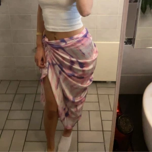 Långkjol i rosa och lila med slits - Säljer en flowy långkjol i mjukt tyg med batikmönster i rosa, lila och vitt. Kjolen har en snygg slits framtill och knytdetalj i sidan som ger en cool omlott-look. Perfekt för sommarens alla häng och vibes. Finns en matchande tröja till i en annan annons.