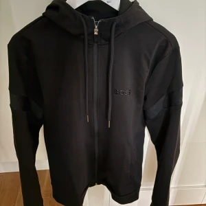 Svart hoodie från BOSS med dragkedja - Svart hoodie från BOSS med snygga detaljer på ärmarna och diskret BOSS-logga på bröstet. Tröjan har huva med snörning, hel dragkedja och långa ärmar. Materialet är mjukt och känns skönt mot huden, perfekt för chill eller när du vill se extra clean ut.