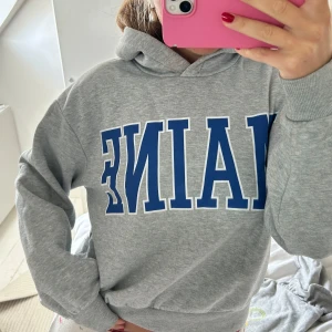 Grå hoodie med blå text - Säljer en grå hoodie med stor blå text på bröstet. Tröjan har huva och ribbade muddar vid ärmslut och nederkant. 🩷