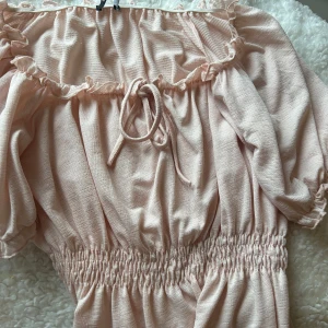 Ljusrosa blus med volang och knyt - Supersöt ljusrosa blus med volangkant och knyt framtill. Smockad midja ger en snygg form och puffiga korta ärmar kan bäras både på axeln och offshoulder. Perfekt för vår och sommar, i mjukt och lätt bomullsmaterial.