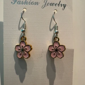 Rosa blomformade örhängen - Superfina örhängen med hängande blommor i ljusrosa och guldfärgad kant. Blommorna har fem kronblad och detaljer i svart, vilket ger en söt och trendig look. Perfekta för att ge din outfit en gullig touch.
